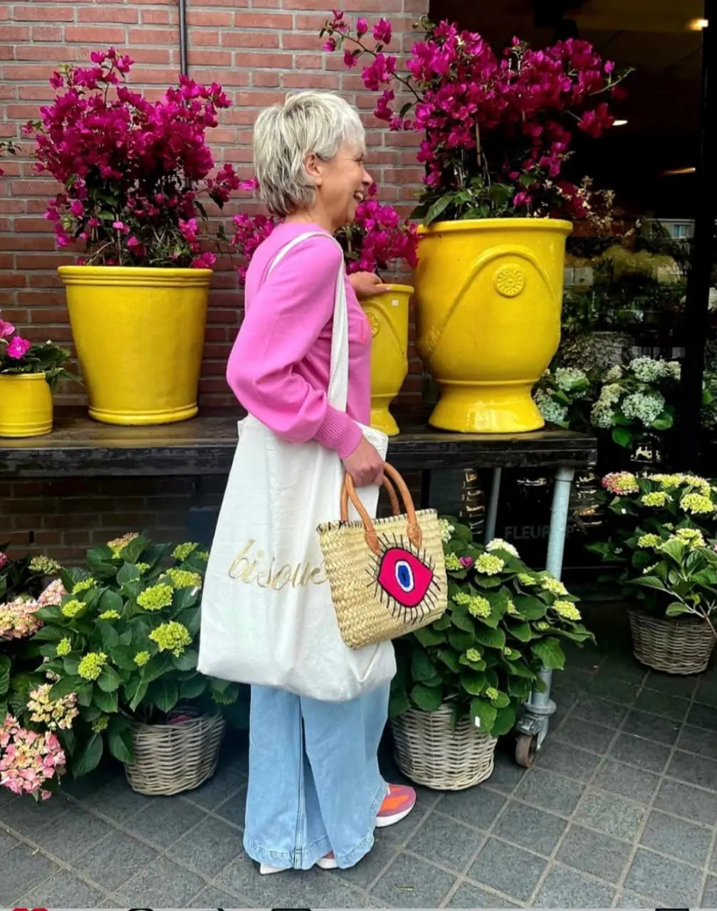 Woman in a pink sweater and canvas tote bag standing next to large yellow flower pots and bougainvillea.