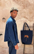Dark denim tote bag with orange palm tree embroidery, displayed beside a model in a denim shirt against a weathered stone wall.