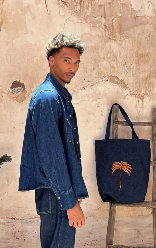 Dark denim tote bag with orange palm tree embroidery, displayed beside a model in a denim shirt against a weathered stone wall.