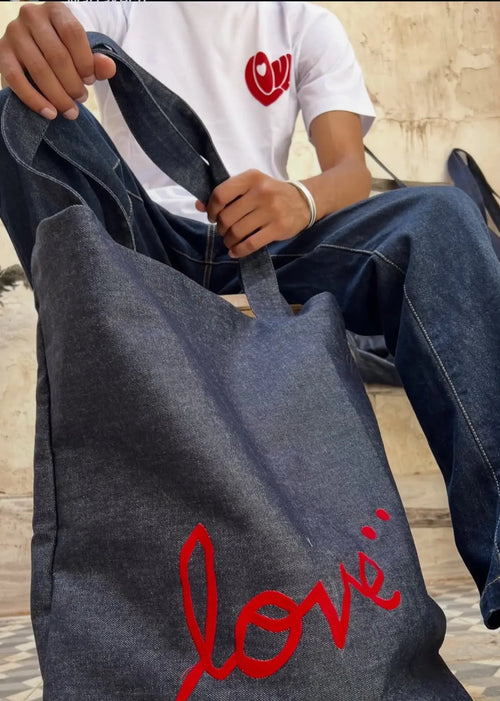 Close-up of a dark denim tote bag with red heart and paint-splatter detailing, held on a model's lap.