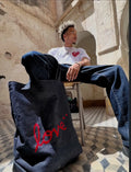 Dark wash denim tote bag with red heart and 'love' script embroidery, held by a model in a white heart-print t-shirt and wide-leg jeans, seated against a weathered stone wall.
