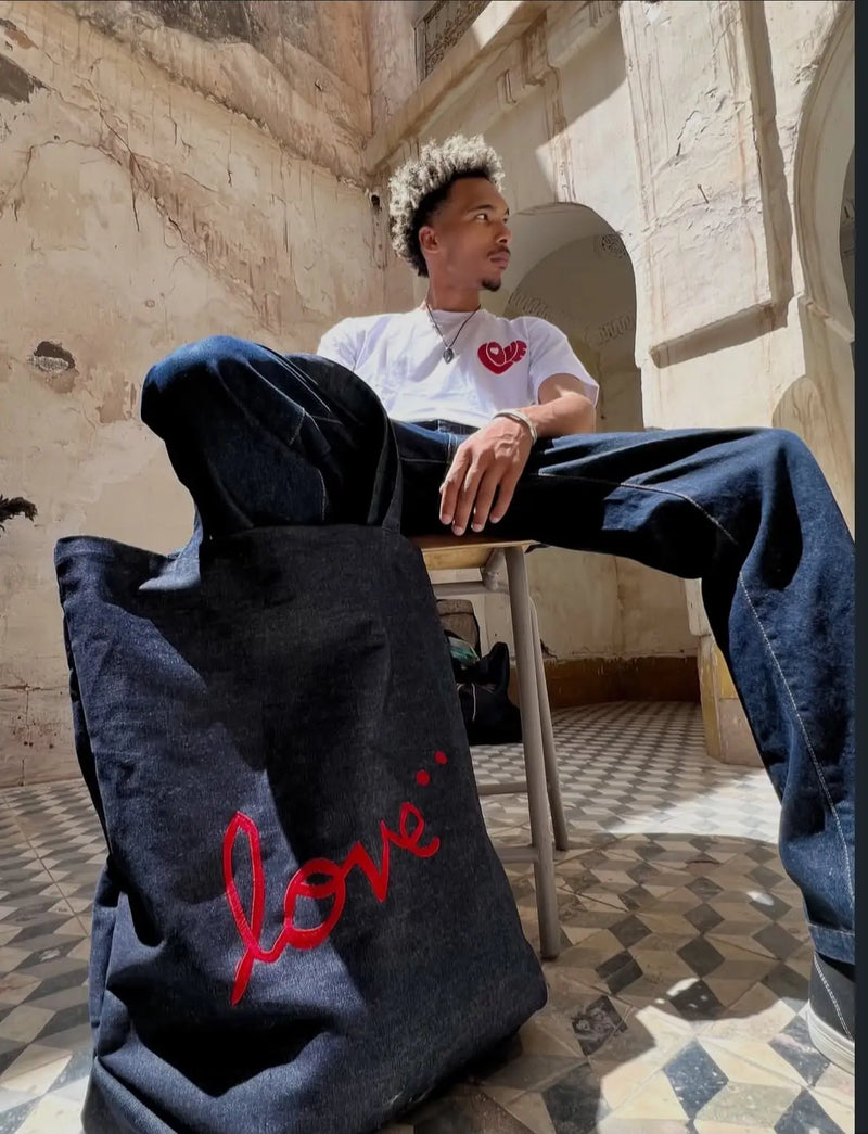Dark wash denim tote bag with red heart and 'love' script embroidery, held by a model in a white heart-print t-shirt and wide-leg jeans, seated against a weathered stone wall.