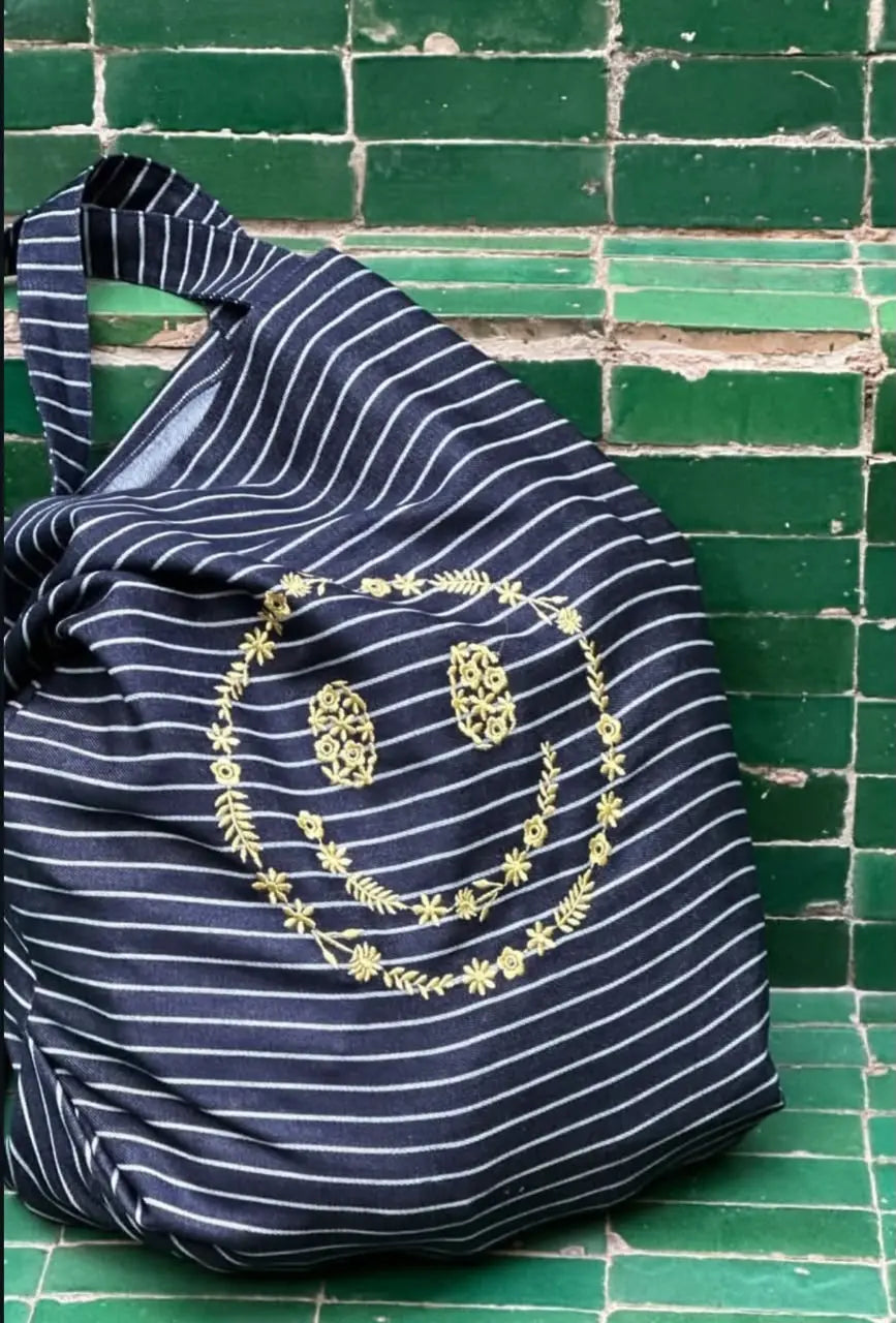 Navy blue striped cloth tote bag with gold floral embroidery detail on green zellige tile backdrop.