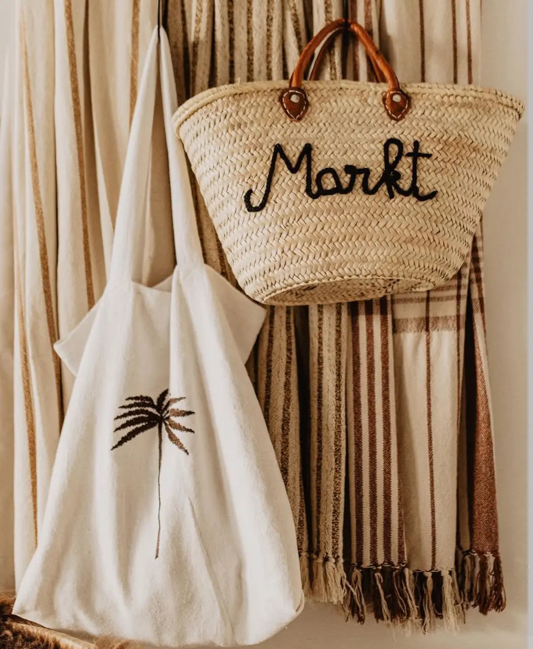 Rustic woven straw market basket bag and embroidered palm tree canvas tote displayed against neutral textured fabric.
